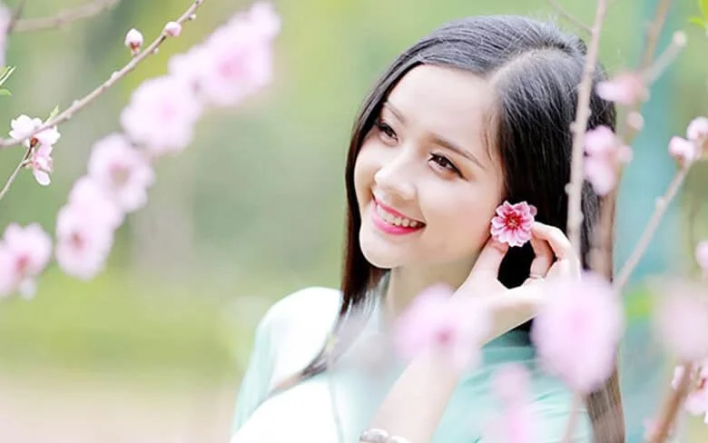 Ảnh cô gái cười tươi bên vườn hoa đào khoe sắc.