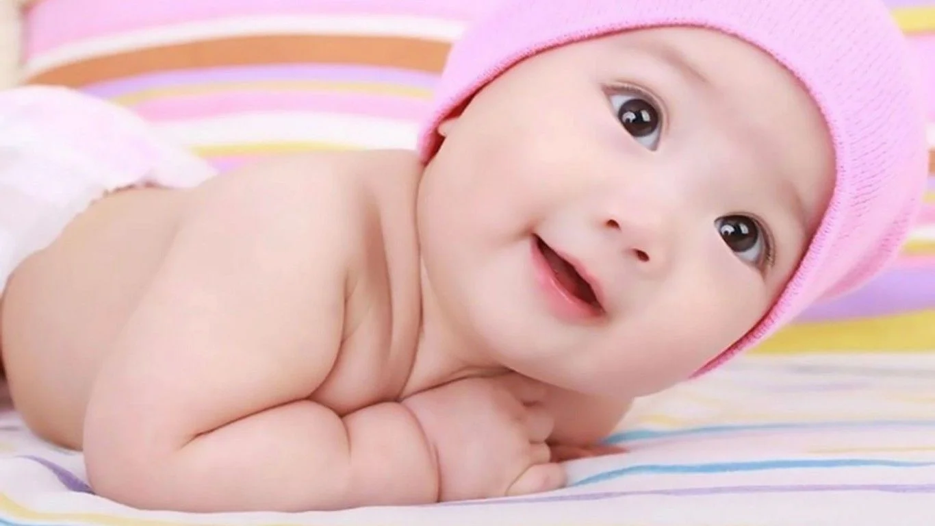 Hình ảnh em bé nằm sấp trên ga giường sọc màu, đội mũ hồng tinh nghịch