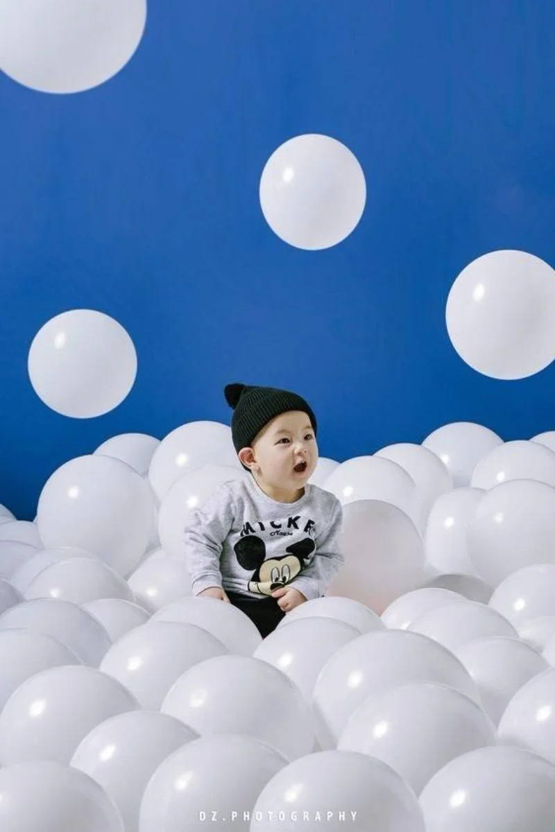 Hình em bé dễ thương đội mũ len đen và mặc áo Mickey Mouse, ngồi giữa những quả bóng bay trắng trên nền xanh dương