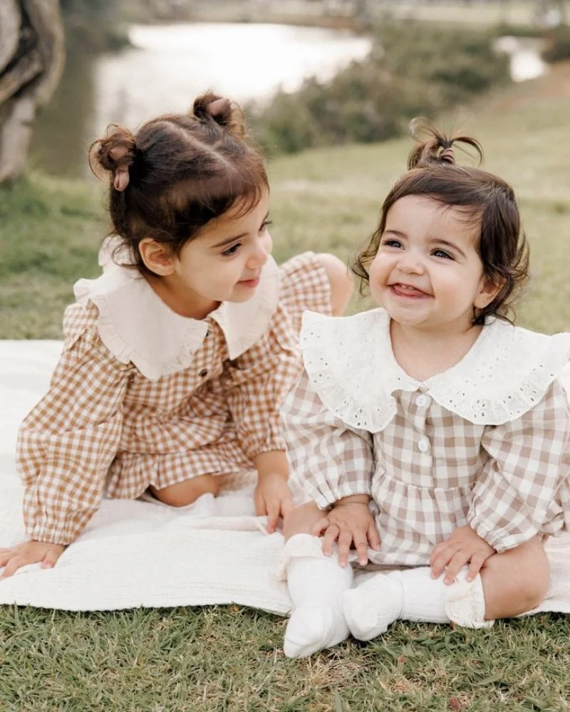 Hai bé gái mặc váy caro cổ sen ngồi chơi trên thảm cỏ, hình em bé dễ thương